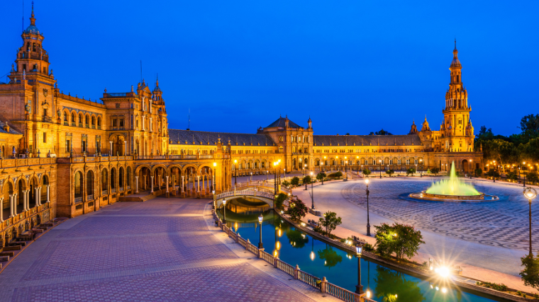 Plaza de España in Seville at dusk, with golden lights illuminating the Renaissance Revival architecture. A central fountain and canal add tranquility.