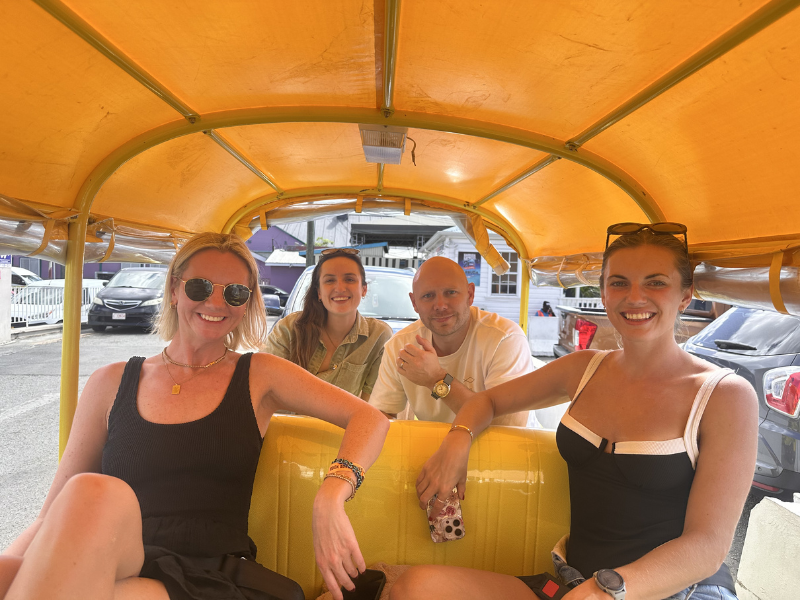 Four people smiling and seated in a bright yellow tuk-tuk with a cheerful atmosphere. The backdrop shows a street scene with cars and buildings in Antigua.
