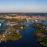 Aerial view of a Oulu city in Finland with numerous buildings surrounded by lush greenery and interconnected waterways under a clear blue sky at dusk.