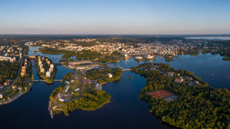 Aerial view of a Oulu city in Finland with numerous buildings surrounded by lush greenery and interconnected waterways under a clear blue sky at dusk.