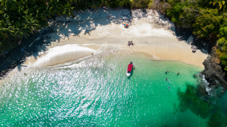 Aerial view of Bolaños, Chiriquí province, Panamá, tropical beach with palm trees, turquoise water, and people swimming near a small boat with a red canopy, evoking a peaceful vibe.