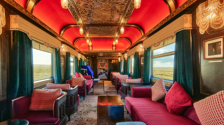 Luxurious vintage train interior on the Golden Eagle Silk Road Express, with plush red sofas, ornate red ceiling, chandeliers, and large windows showcasing scenic countryside views. Elegant ambiance.