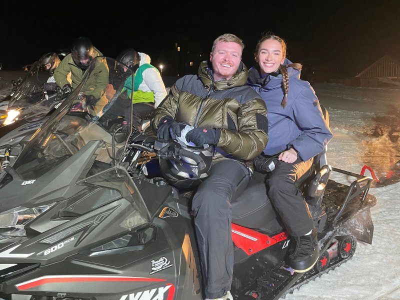 A smiling couple sits on a snowmobile, dressed warmly in jackets under a night sky. Other riders, also bundled up, are visible in the background.