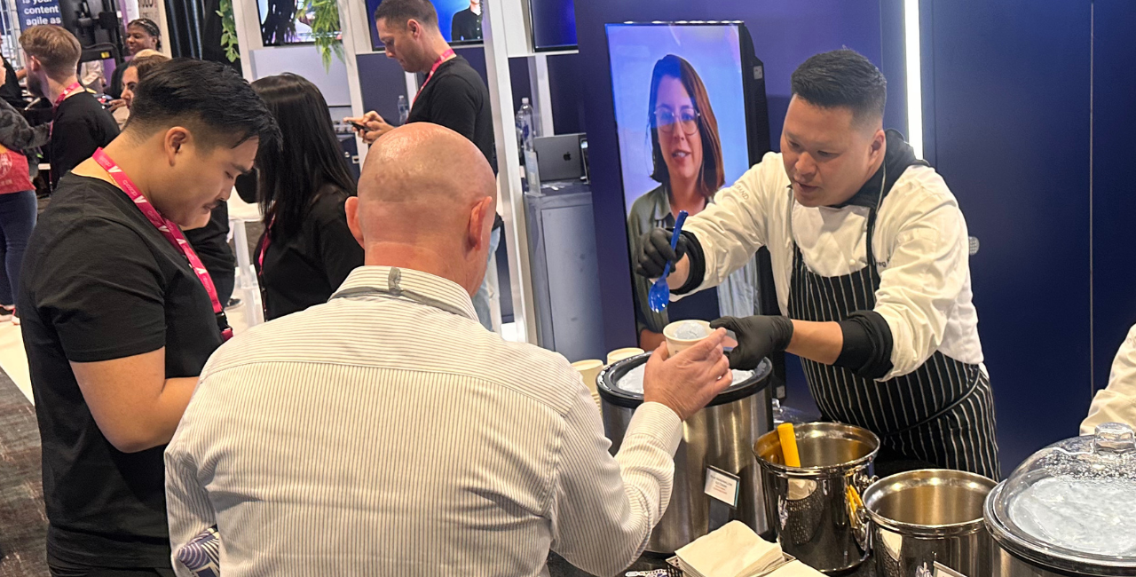 A man in an apron and gloves serves food in cups to two people at a busy indoor event, as attendees enjoying an ice cream on the exhibition stand mingle nearby. A screen behind him displays a woman’s face.