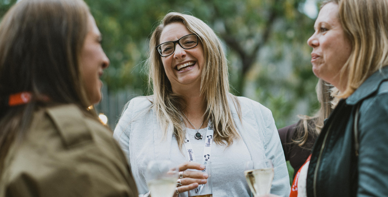 Three women stand outdoors talking and smiling, holding drinks, all wearing event lanyards around their necks, clearly enjoying the experience of connecting at an event.