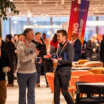 People in business attire converse and network at an indoor corporate event with tables, colourful chairs, and a “TECHNOLOGY” banner in the background.