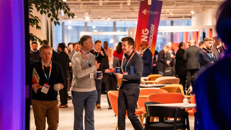 People in business attire converse and network at an indoor corporate event with tables, colourful chairs, and a “TECHNOLOGY” banner in the background.
