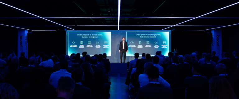 A speaker presents to an audience in a modern conference room with blue lighting, displaying slides titled “Under pressure to change plans, but slow to respond.” The session highlights how activation and event strategy can transform the pace of response during dynamic events.