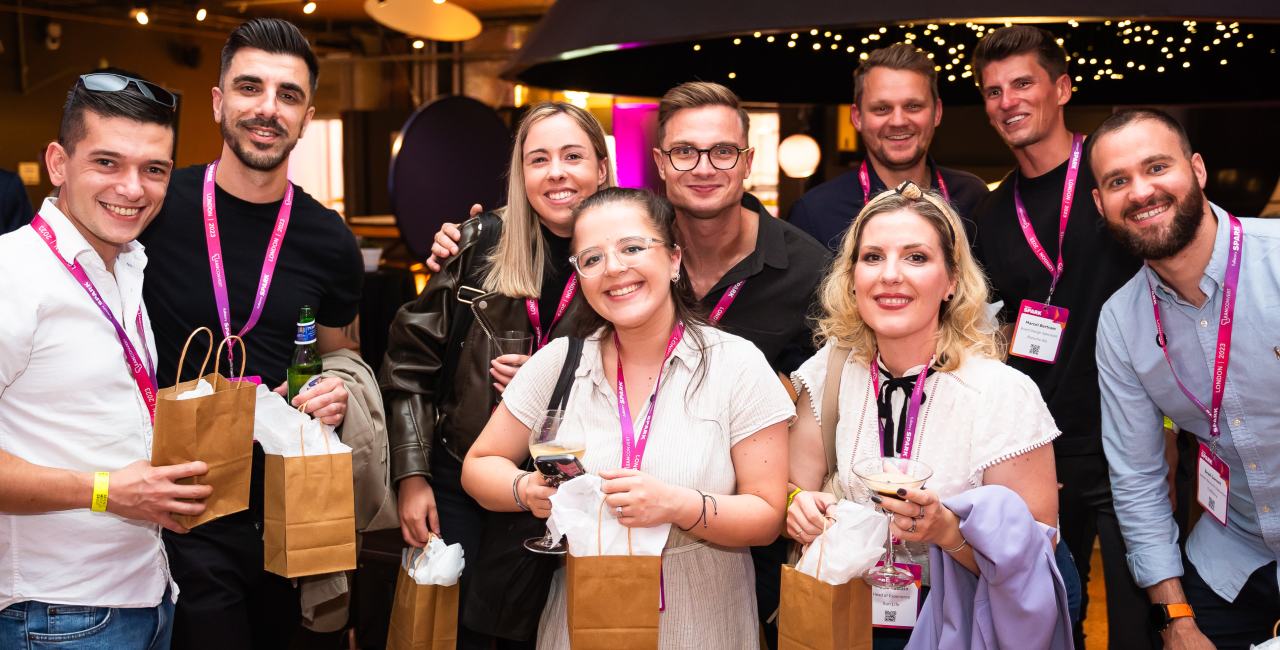 Eight people wearing conference lanyards smile while holding drinks and gift bags at an event.