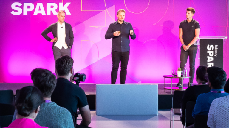 Three speakers on a stage in front of a purple “fullstory SPARK” backdrop, addressing an audience seated in a conference room.
